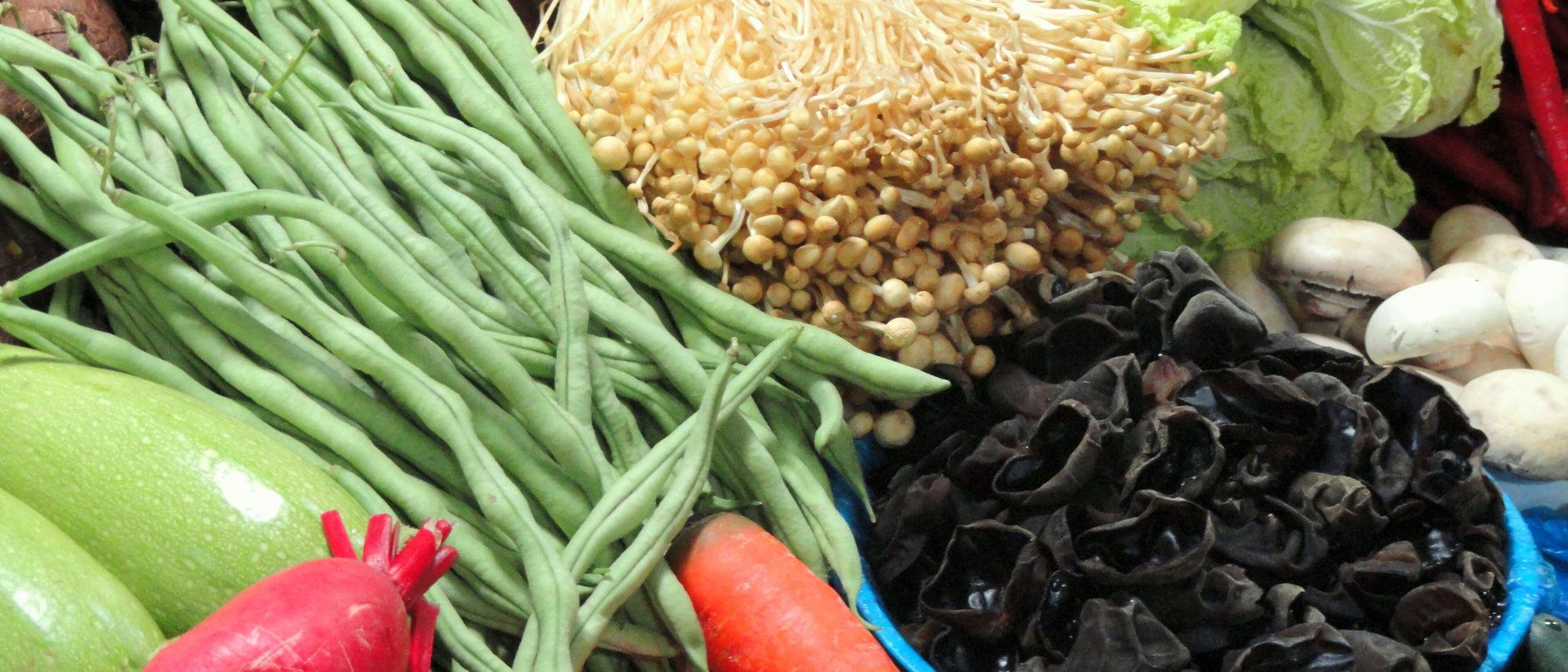 昆明农贸市场一角：翠绿的长豆角、紫茄子、红辣椒与白蘑菇层层叠叠铺满摊位，烟火气十足