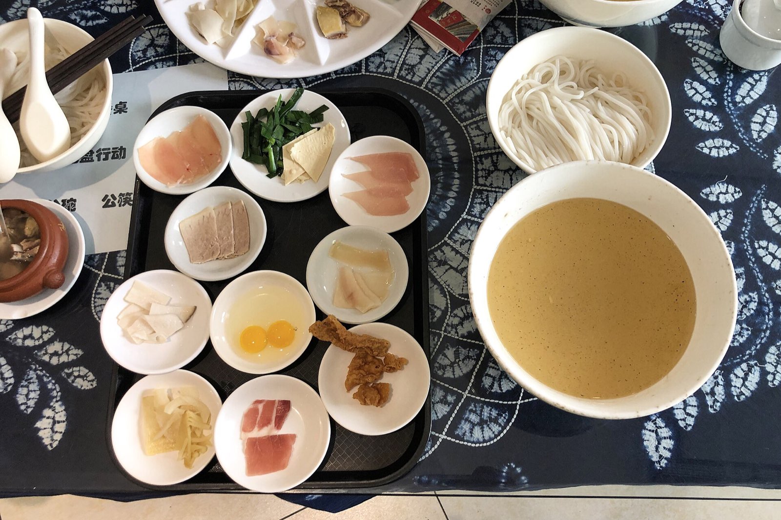 滇式过桥米线的摆盘：黑色托盘里码着肉片、菌菇、双蛋黄等配料小碟，旁边是浓汤与米线