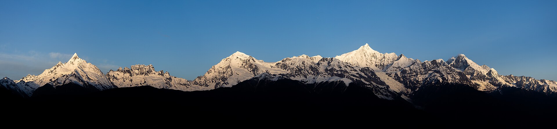 梅里雪山群峰全景：白雪覆盖的山脊在湛蓝天空下连绵起伏，山脚隐入深色剪影